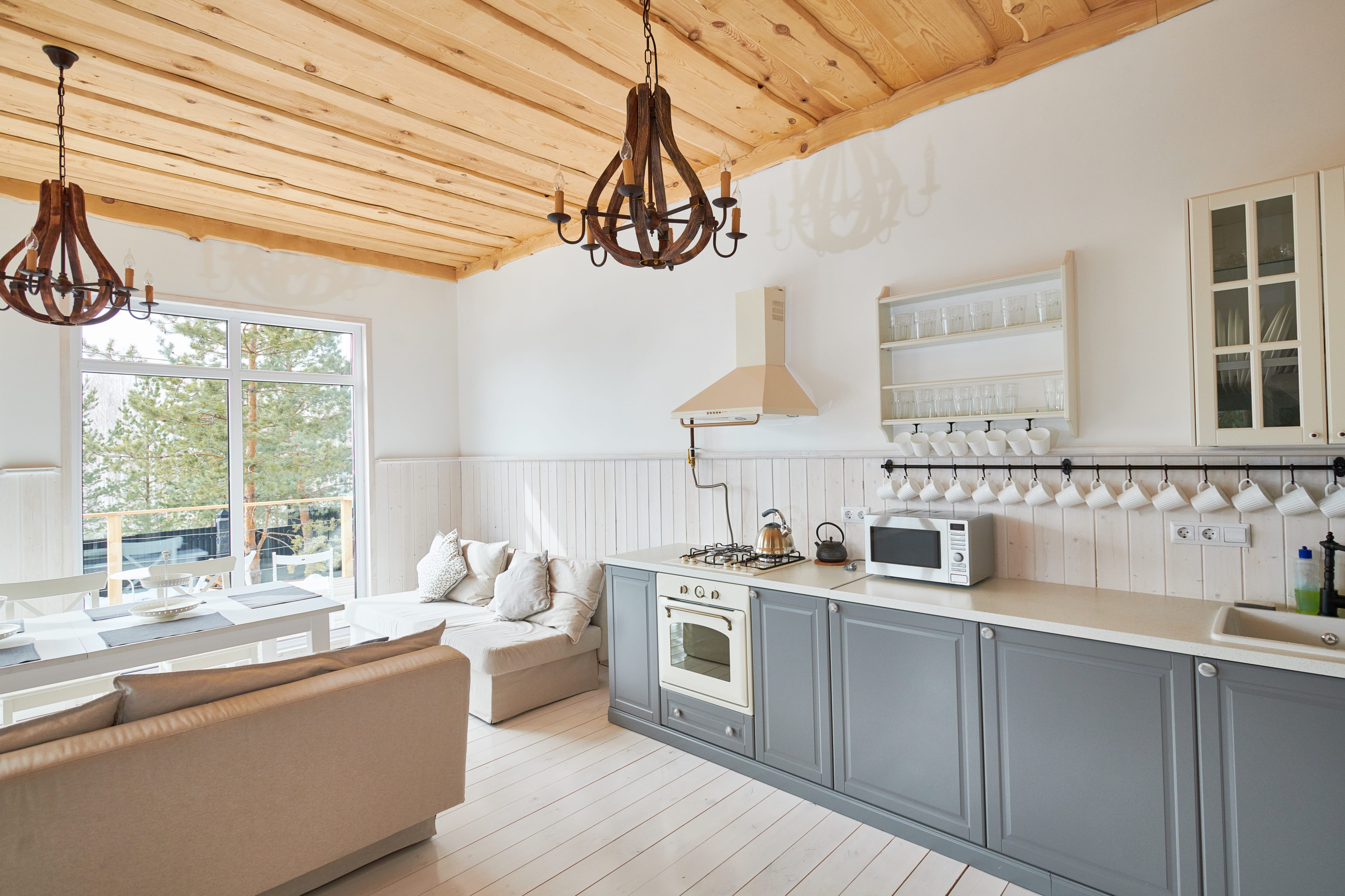 Kitchen with gray cabinets, whitewashed wood walls, 2 iron chandeliers hanging from a wood ceiling, and a nearby dining room 