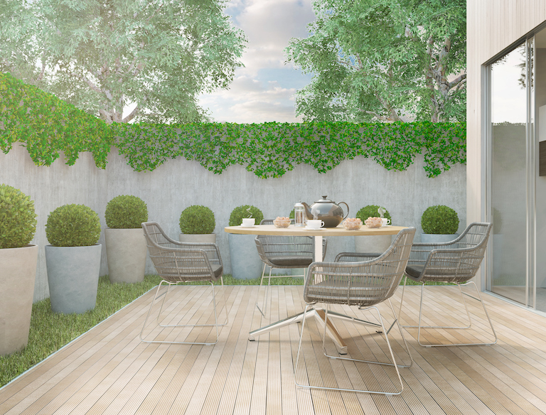light tan deck with green grass and bushes surround it and silver wire deck chairs and table on top
