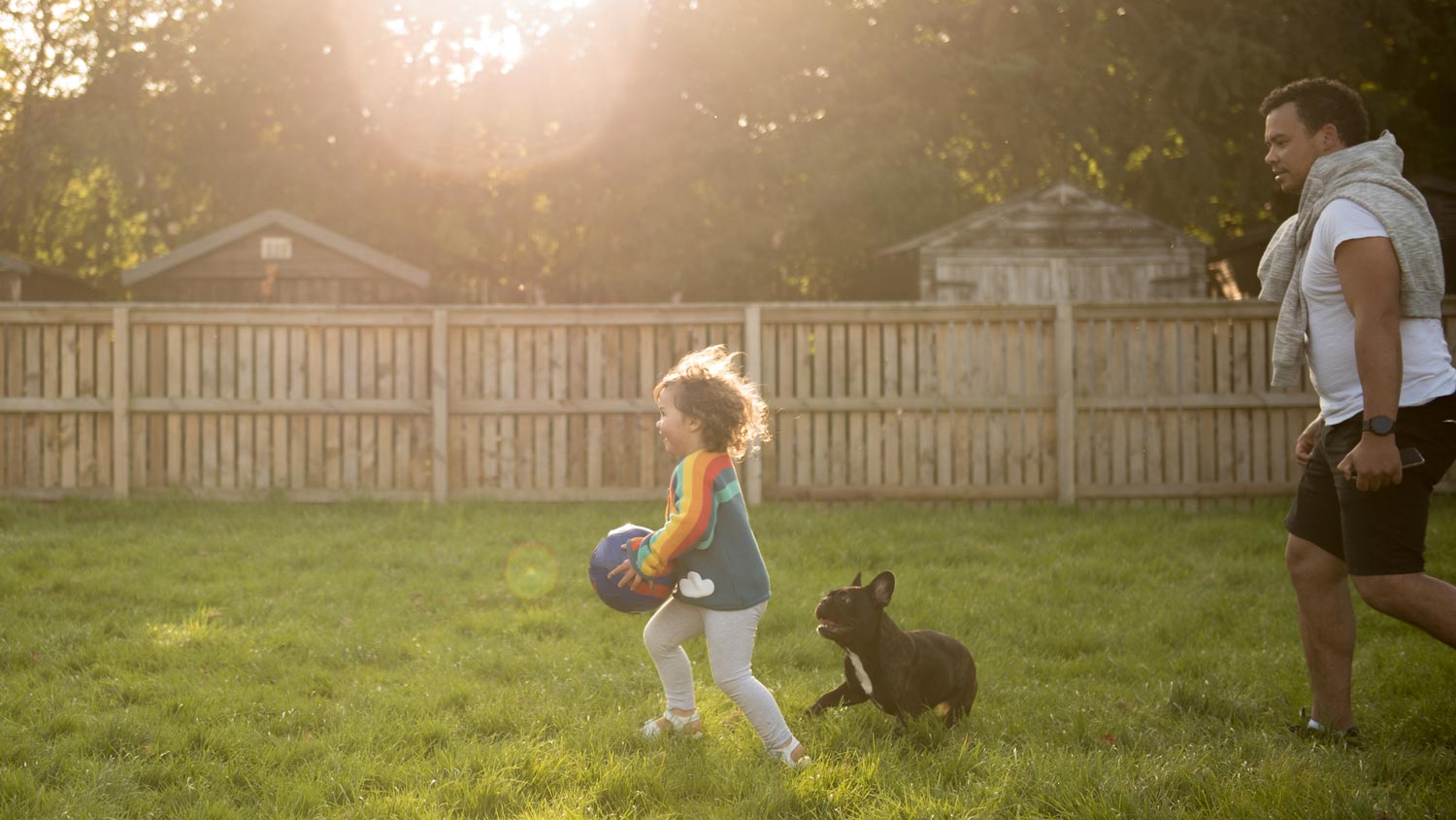 A toddler holding a football running along her father and her dog in the garden